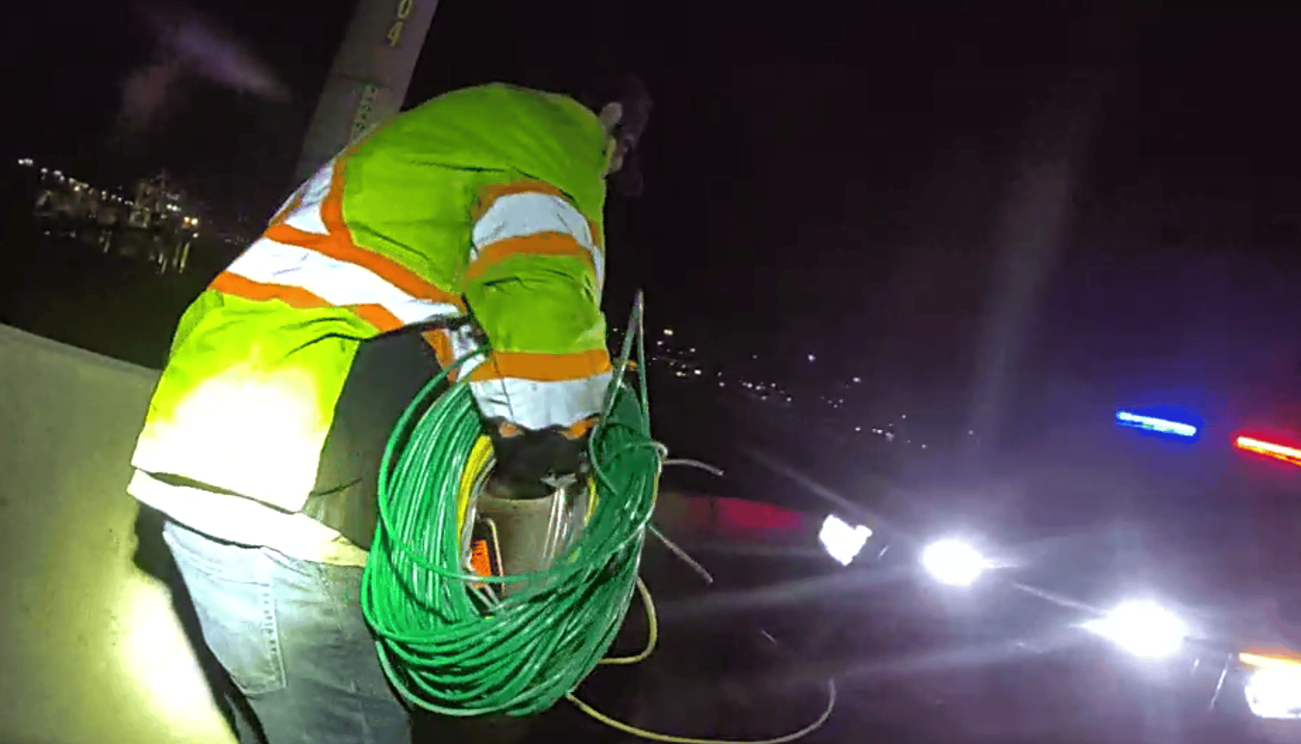 Dark outside early morning. Male suspect wearing reflective jacket, holding green and yellow stolen copper wire. A patrol vehicle parked with headlight on.