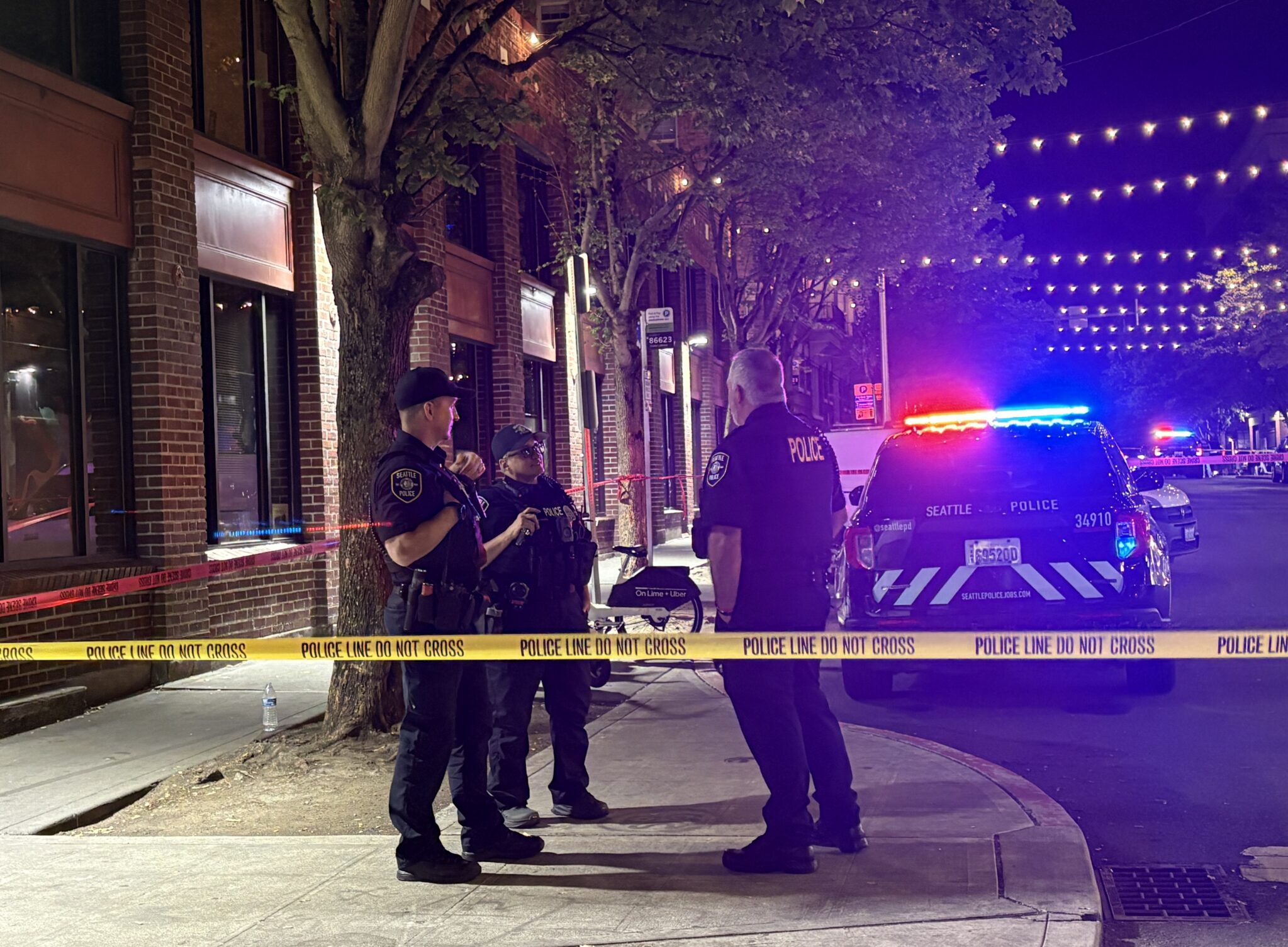 Nighttime picture of three Seattle Police Offices investigating at the scene behind yellow caution tape. a Seattle Police SUV also behind caution tape.